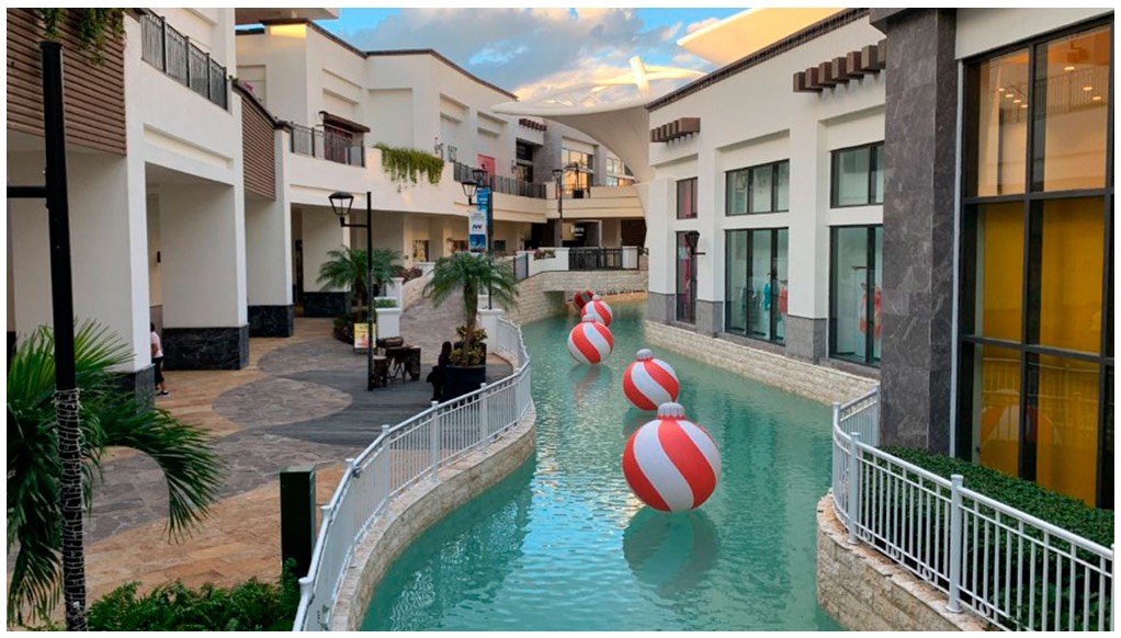 The image shows an artificial river inside a shopping mall. On the river, there are giant spheres. On the sides of the river, you can see commercial premises and vegetation. The image shows a panoramic view of a large area with buildings. Four very large buildings surrounded by vegetation, trees, and pools stand out, giving it the appearance of a residential area. More buildings can be seen on the sides.