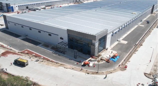 The image is shown in panoramic format and reveals a very large gray building, which appears to be a factory. A large glass entrance and parked vehicles can be seen outside. Behind this esplanade, there is another very long building in the background.