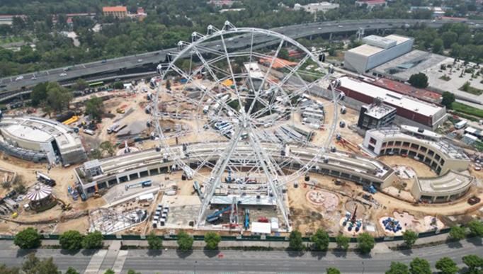 La imagen se muestra en formato panorámico, donde se observa un terreno en el que se está construyendo un parque de diversiones. Se destaca especialmente una gran rueda de la fortuna de dimensiones imponentes